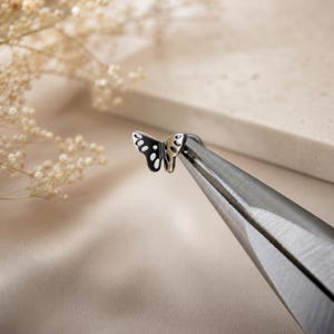 May include: A small, silver-toned metal butterfly charm with cut-out wing details held by a pair of metal pliers. The butterfly features a symmetrical design with open spaces within the wings, showcasing a polished metallic finish and clean edges.