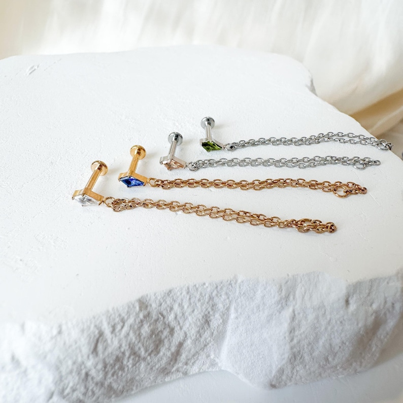 May include: A selection of ear piercing jewellery with chain details. The earrings showcase gemstones in clear, blue, and green, set in gold, silver, and rose gold. The jewellery is presented on a white, textured surface.