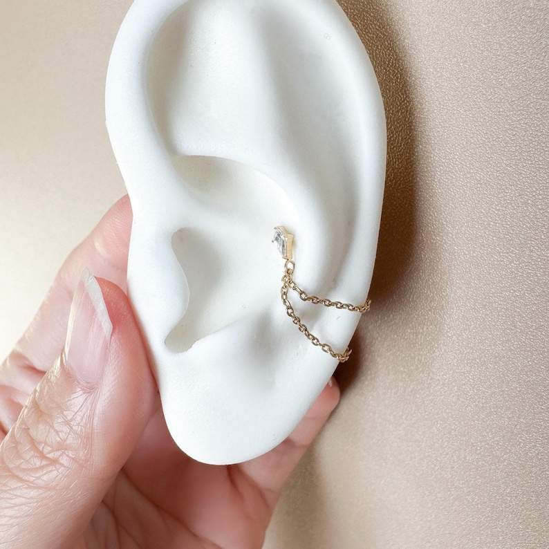 May include: A gold ear cuff featuring a small, clear gemstone and two fine chain strands. The jewellery is displayed on a white ear model, highlighting the design. The background is a neutral beige.