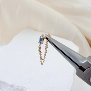 May include: A gold earring featuring a rectangular, pale blue gemstone and a fine chain. The earring is held by pliers, highlighting its design. The earring is set against a white backdrop. The chain hangs down.