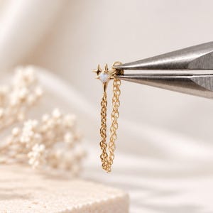 Puede incluir: Un pendiente dorado con un dije en forma de estrella con una piedra iridiscente central y una delicada cadena colgante. El pendiente está sujeto por unos alicates de metal, mostrando el intrincado diseño de estrella y la cadena de eslabones.