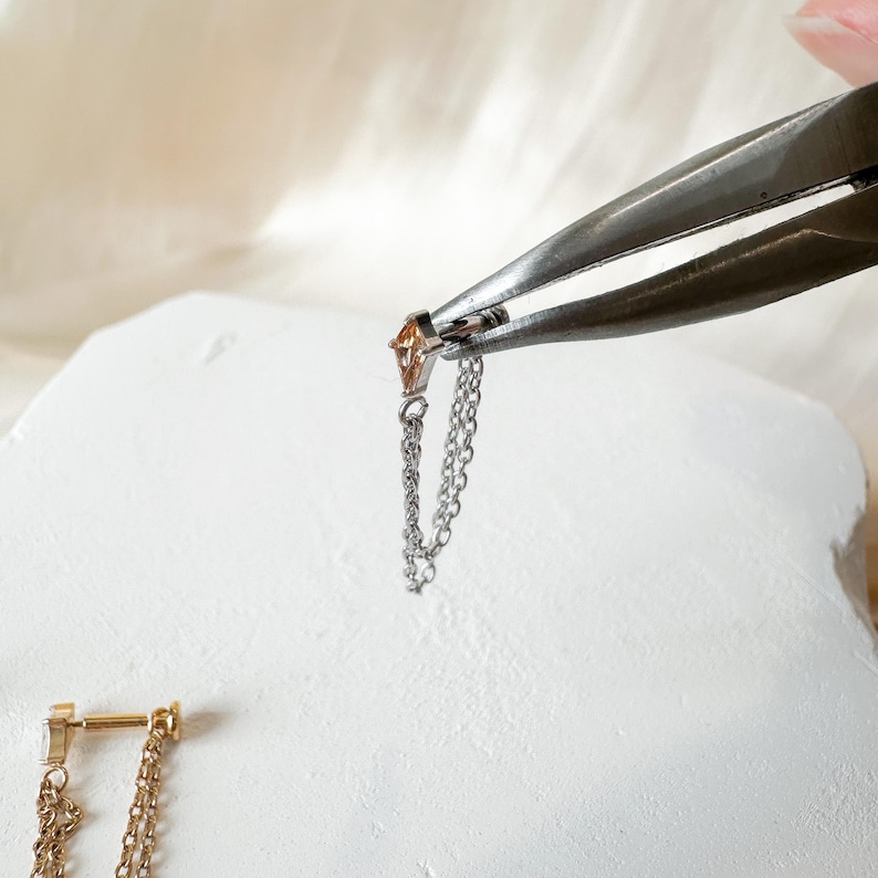 May include: A silver earring with a small, orange gemstone held by pliers. A delicate chain hangs from the earring. Another gold earring with a chain is visible. The earrings are on a white surface.