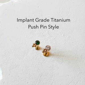 Puede incluir: Dos piezas de joyería corporal de estilo broche de presión de oro con pequeñas cuentas doradas. Una pieza tiene una piedra preciosa verde y la otra una piedra preciosa transparente. El texto "Implant Grade Titanium Push Pin Style" está por encima de la joyería.