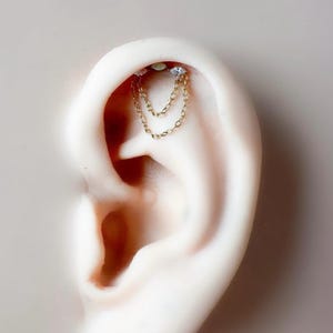 May include: Gold-toned ear jewelry featuring two small, clear gemstones connected by a delicate chain. The chain dangles gracefully, adding a touch of elegance. The jewelry is set against a neutral background, highlighting its intricate design.