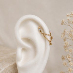 Peut inclure: Un bijou d'oreille doré composé de deux chaînes fines suspendues entre deux points. Une extrémité est ornée d'une petite pierre transparente ronde, tandis que l'autre extrémité et les attaches des chaînes présentent de petites sphères dorées.