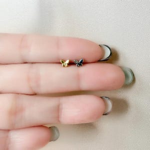 May include: Two small butterfly stud earrings. One is gold-colored, and the other is silver-colored. The earrings are displayed on a light-colored background.