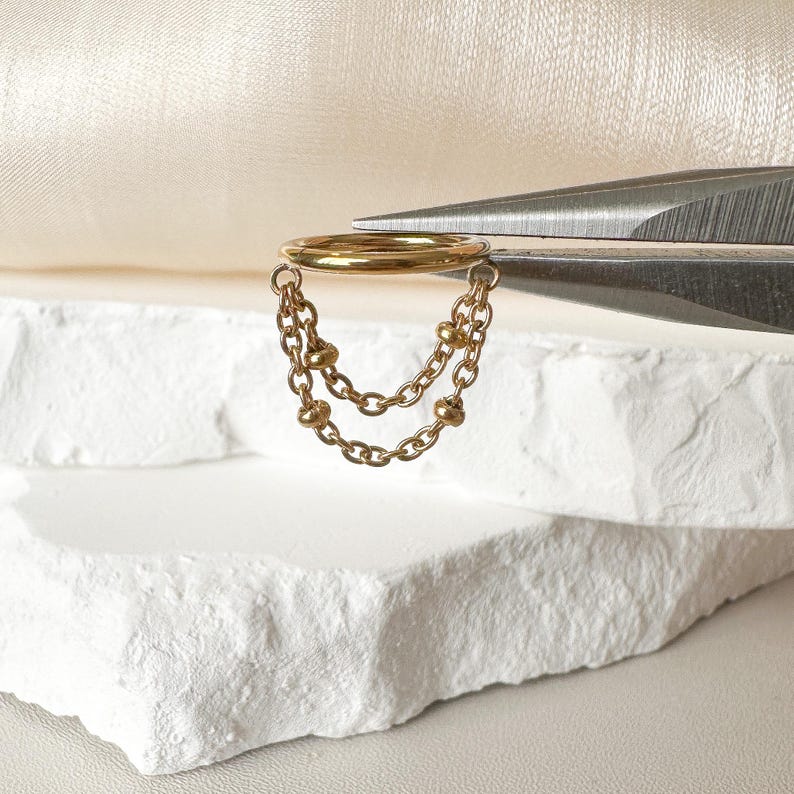 May include: A gold-colored ear cuff with two dangling chains, each featuring small, round beads. The ear cuff is held by a pair of silver scissors against a white and cream-colored backdrop. The jewelry is displayed on a white, textured surface.