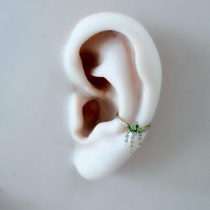 May include: A gold-colored hoop earring with a floral design and dangling charms. The earring features green and blue gemstones. The earring is shown on a white ear model.