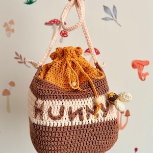 Puede incluir: Bolso de ganchillo hecho a mano en tonos marrón, crema y naranja, con la palabra "HONEY" cosida en la parte delantera. El bolso tiene un cierre de cordón y una pequeña aplicación de abeja. La correa es de color rosa claro.