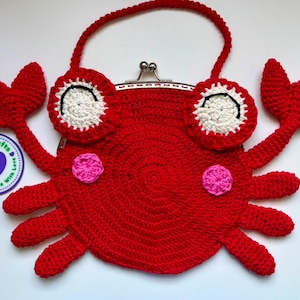 Op de afbeelding: Een felrode gehaakte tas in de vorm van een krab met een metalen knipsluiting. De tas heeft twee wit-rode ogen met zwart stiksel, twee roze ronde wangen en een gehaakt handvat. Een ronde sticker bevat de tekst Dacilipi's Crafts.