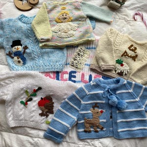 Könnte beinhalten: Eine Sammlung handgestrickter Kinderpullover und -jacken in verschiedenen Farben, darunter Blau, Weiß und Creme. Jedes Kleidungsstück ist mit festlichen Applikationen wie einem Schneemann, einem Engel, einem Rentier und einem Weihnachtspudding versehen. Die Pullover liegen auf einer weißen Oberfläche.