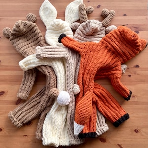 May include: A collection of handmade crochet animal costumes, including a fox, bear, and bunny. The fox costume is orange with black and white accents. The bear and bunny costumes are in shades of brown and cream. The costumes are displayed on a wooden surface.
