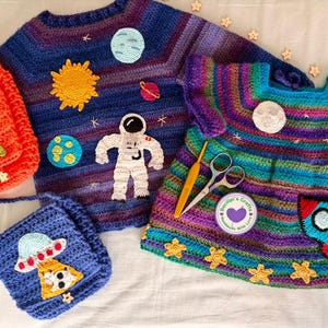 May include: A collection of handmade crochet items with a space theme. Includes a blue and purple sweater with planets and an astronaut, an orange alien bag, a blue UFO bag, and a colorful dress with a rocket and moon.