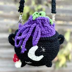 May include: A black crochet cauldron bag with a purple top, featuring a white crescent moon and star embroidery on the front. A small red and white mushroom is attached to the side, and a green frog sits on the rim. It has a black strap.