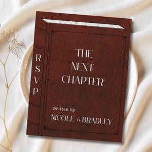May include: A burgundy invitation card designed to resemble a book. The text "THE NEXT CHAPTER" is centered, with "written by NICOLE & BRADLEY" below. The left side reads "RSVP". The card is on a white plate.