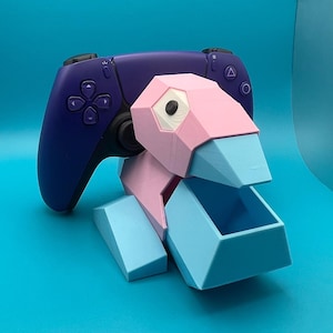 May include: A 3D-printed, geometric Porygon-themed controller stand in pink and light blue, holding a purple game controller. The stand features a box-like structure and a bird-like head, set against a blue backdrop.