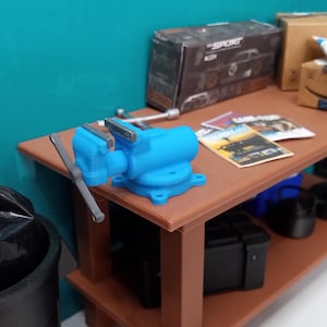 May include: A blue metal vise is clamped to a wooden workbench. The workbench has a lower shelf with a black tool box and a black pot.