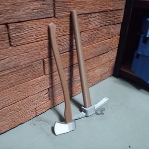 May include: Two miniature tools: a brown-handled axe and a pickaxe. The axe has a silver blade, and the pickaxe has a silver head. Both tools lean against a textured brick wall, suggesting a construction or miniature project theme.