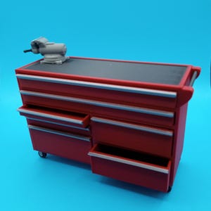 May include: A red tool chest with multiple drawers, some open, and a gray vise on top. The chest has silver trim and sits on small wheels. The background is a solid blue.