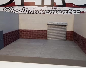May include: A miniature diorama of a garage interior with a brick wall, a closed door, and a roll-up garage door. The diorama is made of cardboard and painted to look like a real garage. The text "@bodymovementrc" is visible on the top of the diorama.
