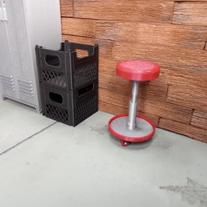 May include: A black plastic crate stacked on another, next to a red and silver rolling stool. A gray metal cabinet and a wood-paneled wall are in the background. The scene is set on a concrete floor.