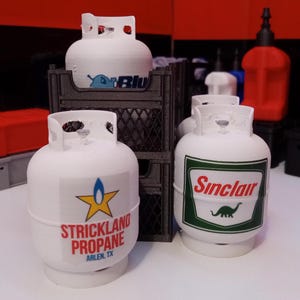 May include: Three white propane tanks with different logos. The tank on the left has a blue and white logo that says "Strickland Propane" and "Arlen, TX". The tank in the middle has a blue and white logo that says "Blue". The tank on the right has a green and red logo that says "Sinclair" and features a green dinosaur.
