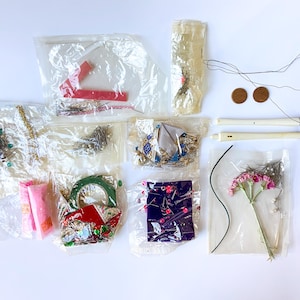 May include: A collection of craft supplies, including pins, beads, fabric scraps, and floral accents, packaged in clear plastic bags. Two small, round, brown objects and two white, cylindrical objects are also visible.