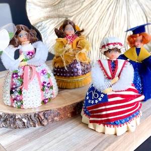 May include: Four vintage doll figurines dressed in different styles. The dolls are made of fabric and sequins and are wearing dresses with various colors and patterns. The dolls are arranged on a wooden surface.
