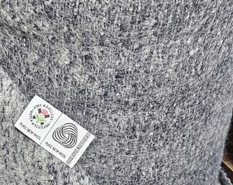 May include: Close-up of a grey woollen textile with a textured surface. A white label with text and a logo is attached to the fabric. The label reads "Pure New Wool" and "Fine Woollens Scotland".