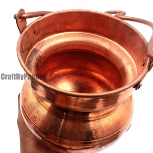 Puede incluir: Una olla de color cobre con base redondeada y boca ancha. Tiene un asa y un asa más pequeña en el lateral. La olla tiene una superficie brillante y reflectante y parece estar hecha de metal. La olla probablemente se utiliza para cocinar o almacenar.