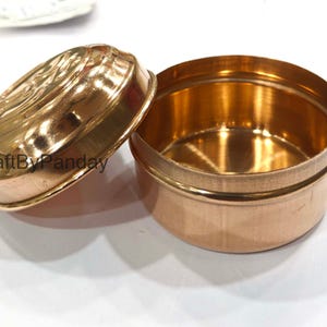May include: A small, open, round, copper-colored container with a matching lid. The lid is slightly ajar, revealing the interior. The lid has a decorative embossed design. The container is likely used for storage.