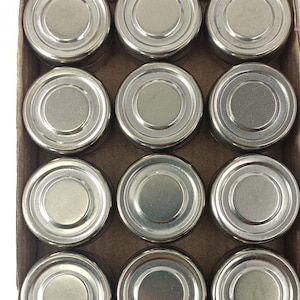 May include: A top-down view of a cardboard box filled with twelve small, round, silver-colored metal containers. Each container has a lid with concentric circular ridges. The containers are neatly arranged in a grid pattern within the box.