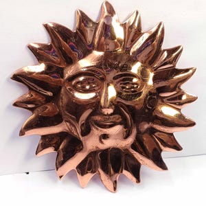 Könnte beinhalten: Kupferfarbene Sonnen-Wanddekoration mit Gesicht und stacheligen Strahlen. Die Sonne hat oben ein Loch zum Aufhängen. Das Metall hat eine glänzende, reflektierende Oberfläche.