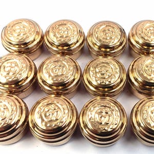 Puede incluir: Doce pequeños recipientes redondos de color dorado con tapas. Cada tapa presenta un diseño en relieve detallado. Los recipientes están dispuestos en tres filas de cuatro, mostrando su apariencia uniforme y brillo metálico.
