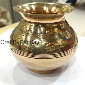 May include: A small, round, brass-coloured pot with a wide, flared rim. The pot has horizontal etched lines and a slightly bulbous body. The text "CraftByPanday" is visible on the side of the pot.