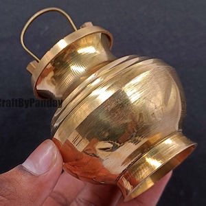 Könnte beinhalten: Ein kleiner, verzierter Messingtopf mit Henkel. Der Topf hat einen abgerundeten Körper mit ausgestelltem Boden und Deckel. Der Henkel ist eine einfache Schlaufe. Der Topf wird in einer Hand gehalten und scheint poliert zu sein.