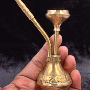 May include: A brass smoking pipe with a curved stem and a textured mouthpiece. The pipe has a bulbous base with an engraved floral design. The pipe is held in a hand against a dark background.
