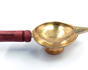 Peut inclure: Une lampe à huile en laiton avec une poignée en bois. La lampe a une forme de bol rond avec un bec pointu et une base courte et ronde. La poignée est de couleur rouge foncé et le texte "CraftByPanday" est imprimé sur la poignée.