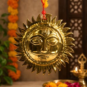 May include: A polished brass sun wall hanging featuring a stylized face with a mustache and a forehead mark. The circular metal plaque has radiating pointed rays and is suspended by a thin red string with a small clear bead at the top.