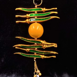 Puede incluir: Un colgante dorado con detalles verdes y amarillos. Presenta formas doradas curvas con líneas verdes, una esfera amarilla central y una cadena con pequeñas cuentas. El fondo negro resalta el diseño intrincado.