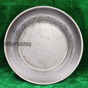 Könnte beinhalten: Eine runde, silberfarbene Metallpfanne mit leicht erhöhtem Rand. Die Pfanne hat ein abgenutztes Aussehen mit dunklen Flecken und Kratzern. Der Text "ftByPanday" ist auf der Seite der Pfanne aufgedruckt. Die Pfanne steht vor einem grünen Hintergrund.
