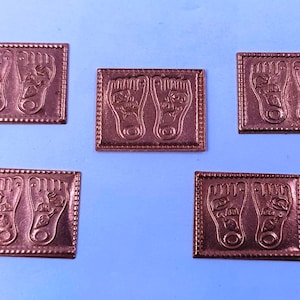 Puede incluir: Cinco baldosas cuadradas de color cobre con un diseño en relieve de dos pies. Cada baldosa presenta un contorno detallado de los pies, incluyendo dedos y plantas. Las baldosas están dispuestas sobre una superficie azul claro.