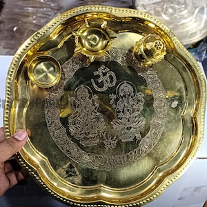 May include: A gold-colored metal puja thali with a scalloped edge. The thali features engraved images of deities, the Om symbol, and other religious inscriptions. It includes small bowls and a lamp holder.