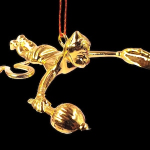 May include: A gold-colored ornament in the shape of a stylized figure, possibly a monkey or a similar creature, with outstretched arms and a bulbous object in one hand. The ornament is suspended by a red string against a black background.
