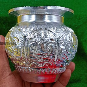Può includere: Un vaso argentato e ornato con intricati disegni in rilievo. Il vaso ha un'ampia bocca e un corpo arrotondato, tenuto in mano su uno sfondo verde. Le sculture dettagliate presentano motivi floreali e forse animali.