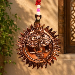 May include: A polished copper sun wall hanging featuring a stylized face with a mustache and radiating sunbeams. The circular metal piece is suspended by a red cord adorned with two white beads and one central pink bead at the top attachment point.
