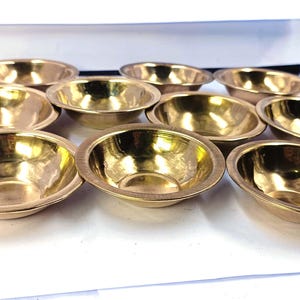 May include: Eleven small, oval brass bowls with a shiny, golden finish. The bowls are arranged on a white surface. These bowls are likely for serving or decorative use, and are made of brass.