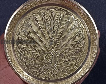 Op de afbeelding: Een decoratieve messing plaat met een reliëf pauwenontwerp. De veren van de pauw zijn uitgespreid en de plaat heeft een gedetailleerde bloemenrand. De plaat is rond en goudkleurig.