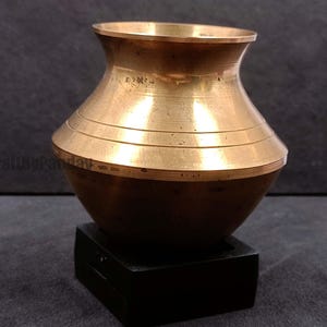 May include: A small, antique brass pot with a rounded base and a flared rim. The pot features horizontal bands around the middle and sits on a black square base. The brass has a warm, golden hue.
