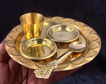May include: A golden brass puja set, including a small plate with a scalloped edge, two small bowls, a small cup, and a spoon. The set is designed for religious rituals and ceremonies.